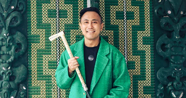 Chris Tse, smiling and wearing a green suit, holding a carved tokotoko and standing in front of a green and beige tukutuku panel.