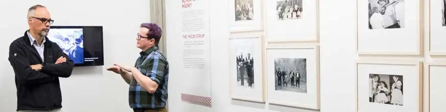Two men standing and talking in front of the images from the Māori Strum section of the Pūkana exhibition.