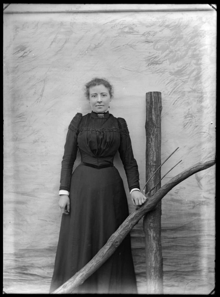 A woman in dark-coloured dress stands beside a pitchfork in a posed scene in front of a painted backdrop.