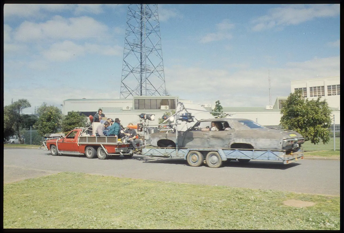 Film crew sitting on the back of a ute filming people in another old battered car being pulled on a rig behind them.