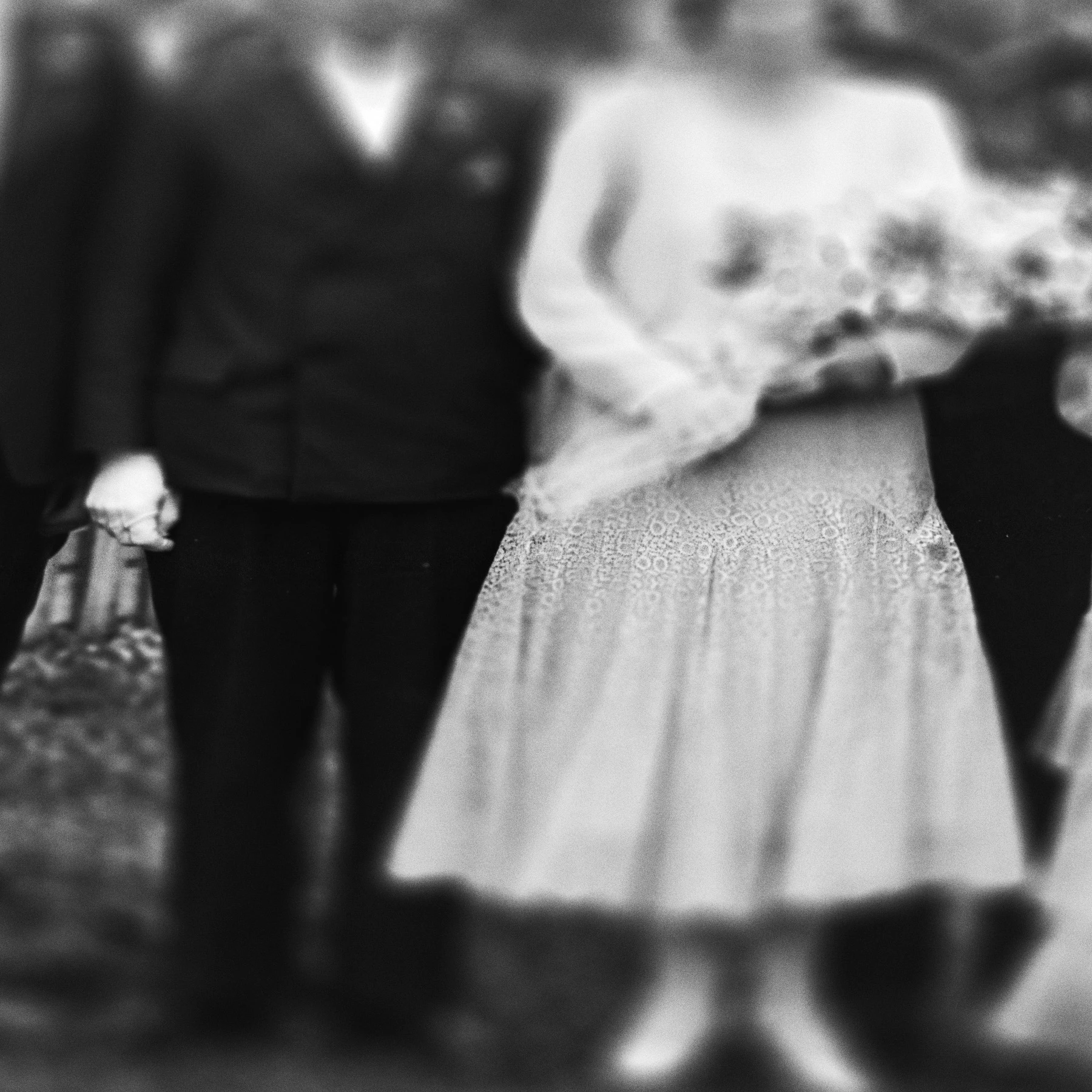 A man and woman stand together in formal wedding attire, she is holding flowers but both their heads and feet are blurry.