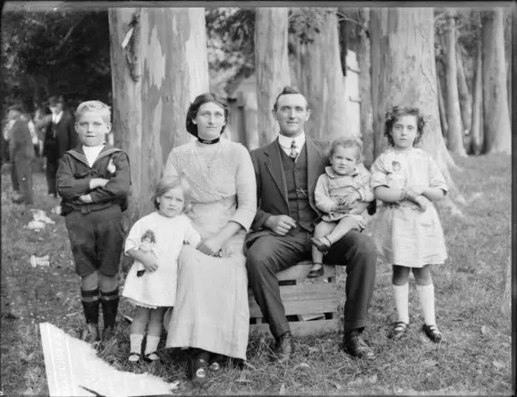 Family portrait under gum trees, unidentified parents with young son and two daughters with dolls, baby girl on father's knee, wooden building and people beyond.