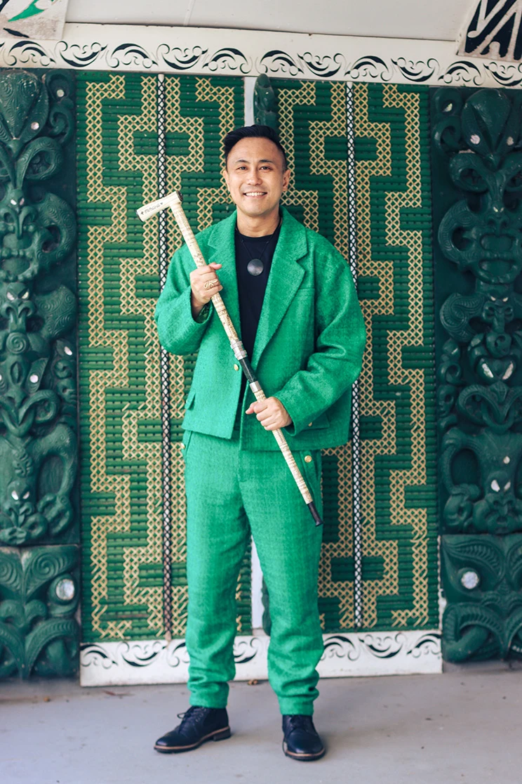 Chris Tse, smiling and wearing a green suit, holding a carved tokotoko and standing in front of a green and beige tukutuku panel.