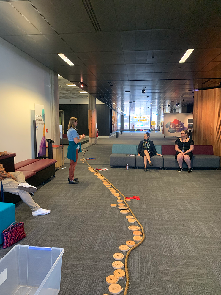 A woman standing talking to people who are sitting. There is a long rope on the ground with small wooden panels hanging off it. 