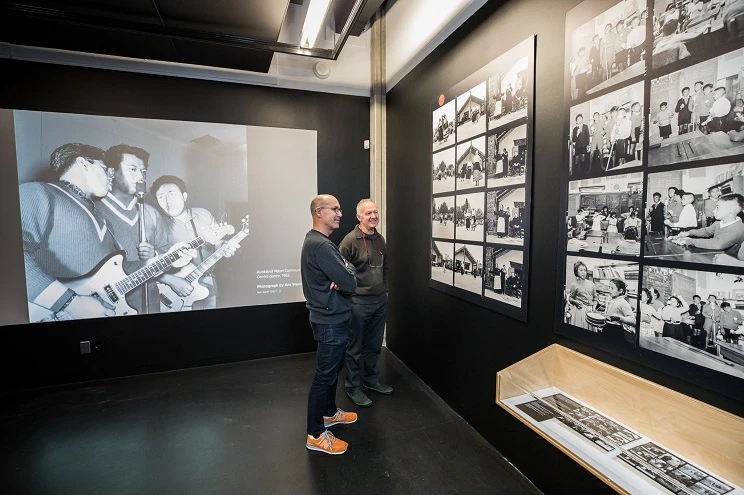 A scene from inside a gallery with the lights turned on, showing two men standing in front of a wall looking at the enlarged contact sheets which have been hung on it, and a large black and white photo projected on the adjacent wall.