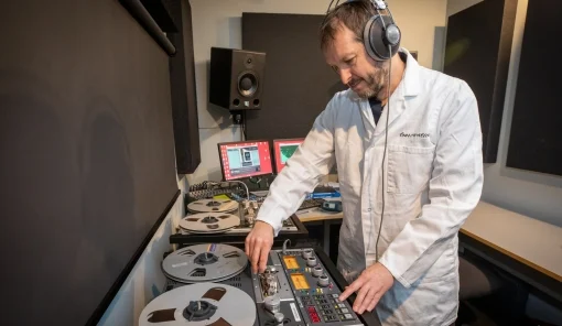 AV technician seen in white lab coat operating a large reel to reel tape machine while wearing headphones. 