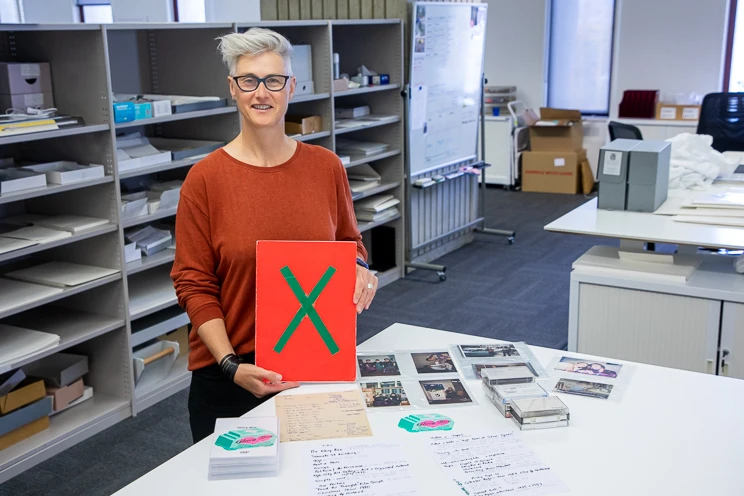 A woman stands behind a table on which sit an array of paper documents, cassettes and photos, and in her hands she holds a large red booklet with a green 'x' on the cover.