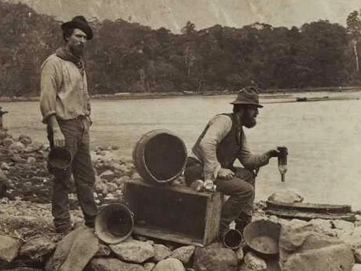 Two men beside a lake with empty buckets, one man holds a bottle upside down to show it contains no contents.