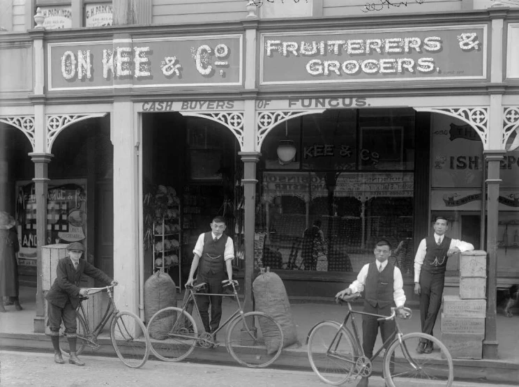 Four men stand in front of a grocery shop with three holding bicycles.