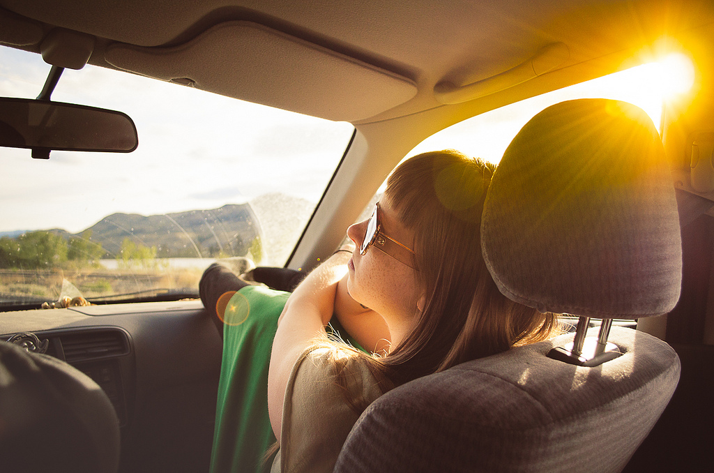 A girl wearing sunglasses sitting in the front seat of a car. She has her feet on the dashboard. The sun is shining through the window.