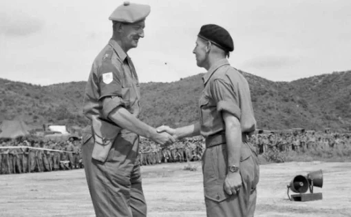 Two men in military dress shaking hands in front of a crowd of fellow soldiers.