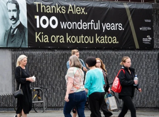 Pedestrians crossing the street beneath a banner with the words, 'Thanks Alex, 100 wonderful years. Kia ora koutou katoa', alongside a picture of Alexander Turnbull. 