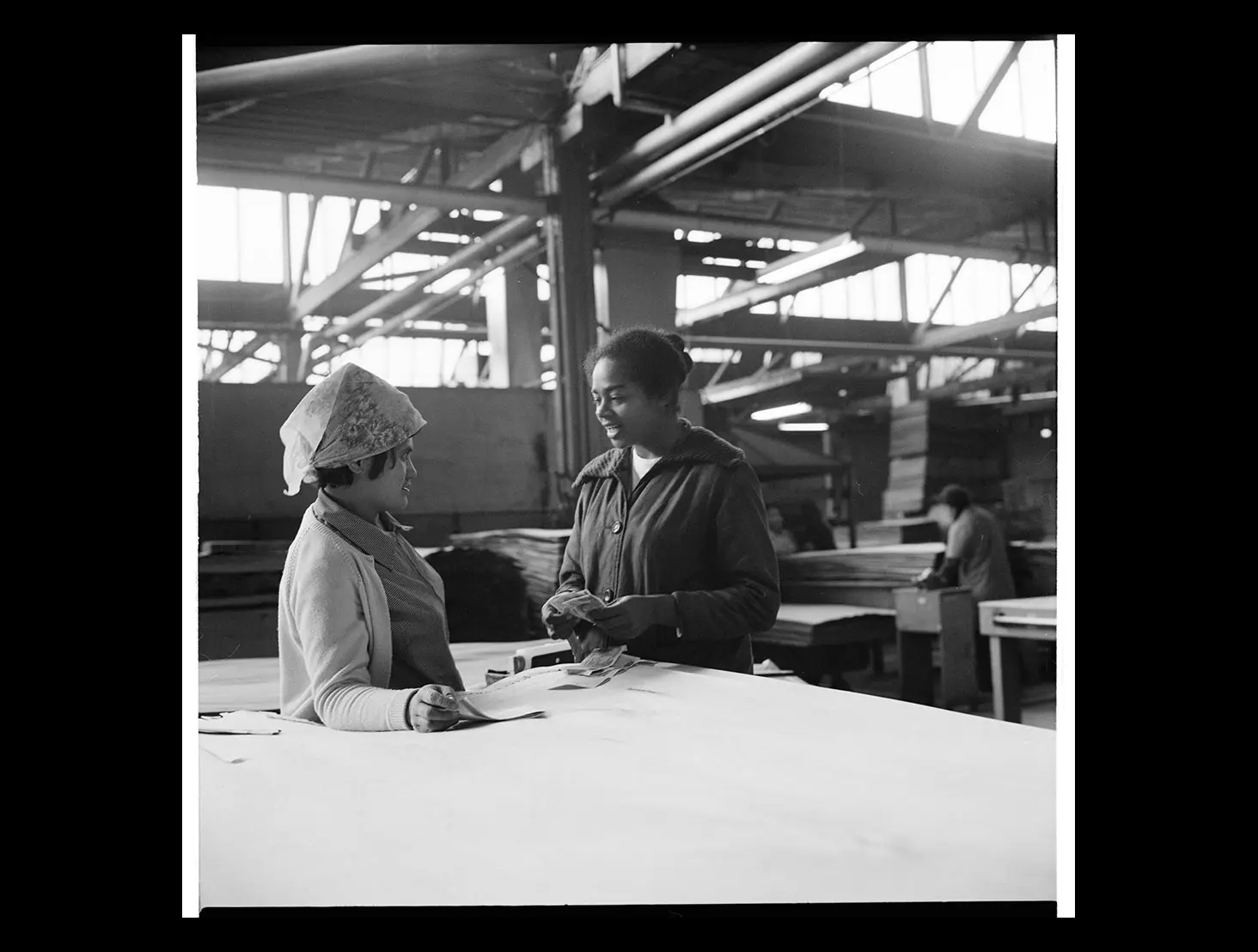 Black-and-white photo of 2 female Pacific Island workers talking in a plywood factory.