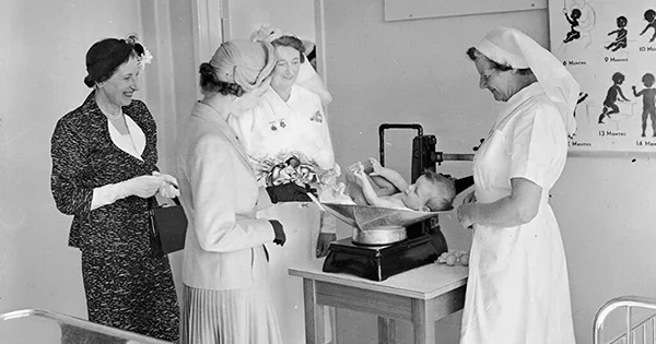 Two nurses and two women wearing skirt suits, hats and gloves smiling at a baby lying on baby scales set on a table.