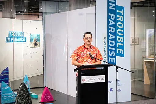 Man standing at podium talking. He is wearing a colourful shirt with flowers on it. Behind him are the words Trouble in Paradise. 
