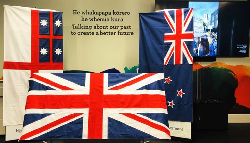 3 flags of Aotearoa New Zealand displayed in the He Tohu Tāmaki exhibition space in front of words:
'He whakapapa kōrero he whenua kura. Talking about our past to create a better future'.