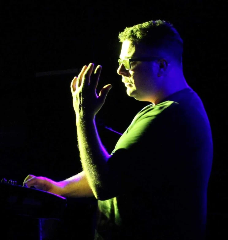 A man wearing glasses performs on stage, backlit with a blue light and front lit by yellow/green light.