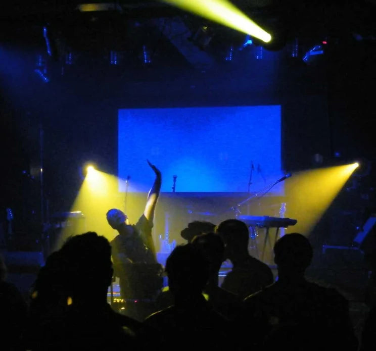 A view of the stage (empty) with dramatic yellow and blue lighting and members of the crowd silhouetted.