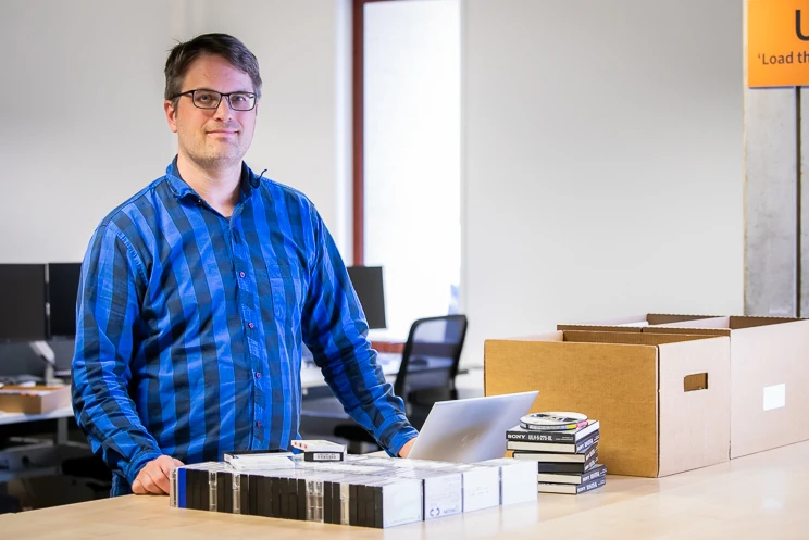 A man stands at a table with a laptop open and a large stack of cassettes neatly arrayed in front of him, along with a few open reel tapes.