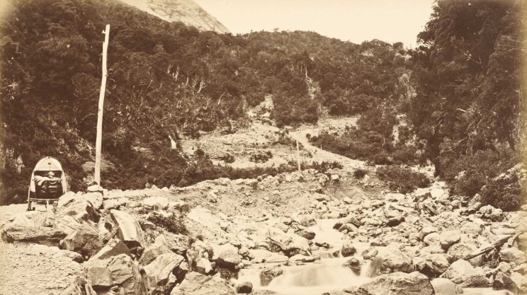 Rugged and rocky road beside a creek that leads up and over a pass in the distance. A buggy is on the side of the road on the left of the image.
