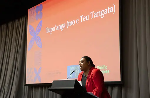 Tongan woman at lectern speaking. Behind here is a screen with words "Tupu'anga (mo e Teu Tangata).