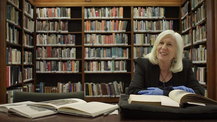 Woman sitting at a desk in front of lots of bookshelves, wearing gloves and looking at heritage books. 