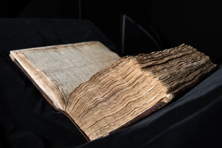 A three-quarter angle shot of a large, bound volume which sits open halfway, its uneven, yellowing pages clearly visible in the dramatic lighting.