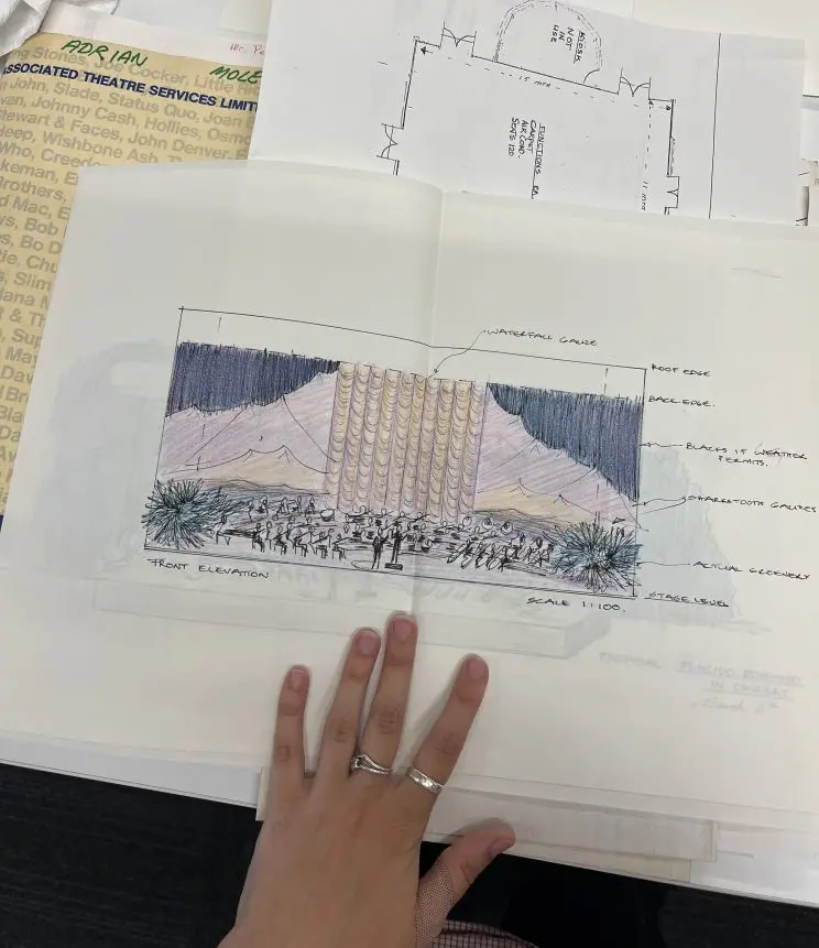 On a table is an array of drawings, elevations and schematics that depict the stage design for an orchestra complete with 'waterfall gauze' behind the orchestra at the back of the stage.