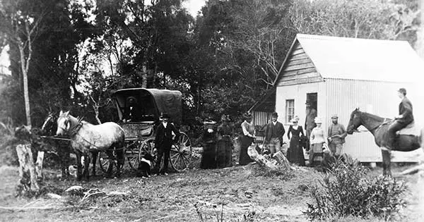 Black and white photo of a group of men and women, a horse and carriage, and a man on a horse outside a small building.