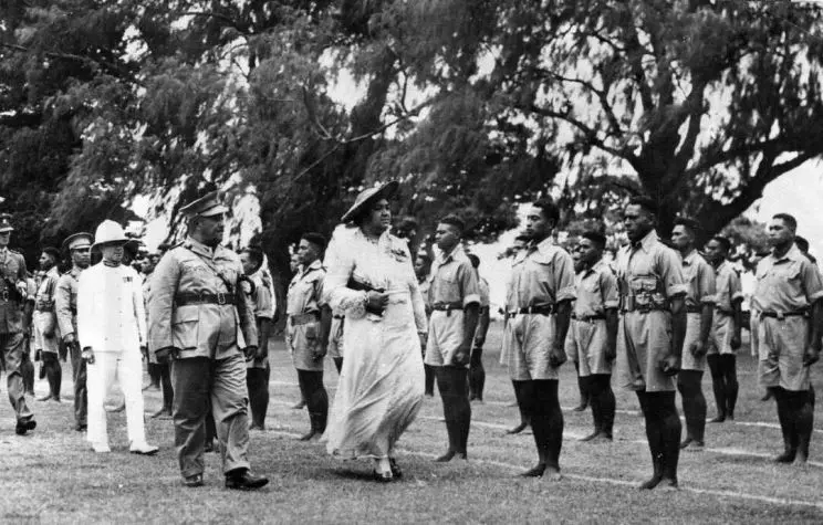 A woman in formal dress can be seen being given a tour by a man dressed in military uniform alongside troops standing at attention. 