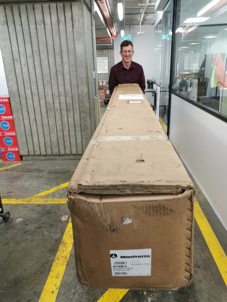 A very large, long cardboard box has arrived at the Library and staff member is taking it to the studio.