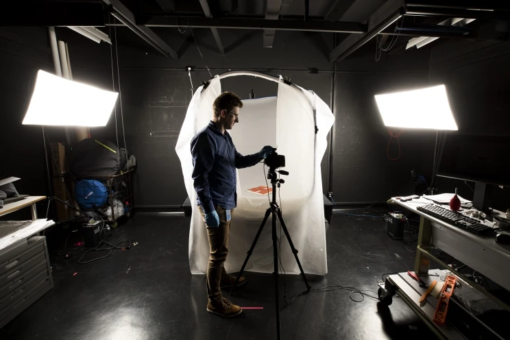 Photographer stands in the studio with camera on tripod and special lights illuminating the subject hidden inside a light tent.