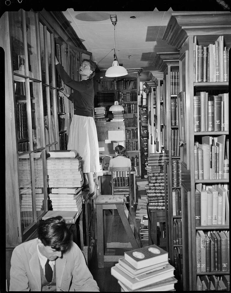 Floor to ceiling bookshelves, men and women librarians reading and looking at the shelves.