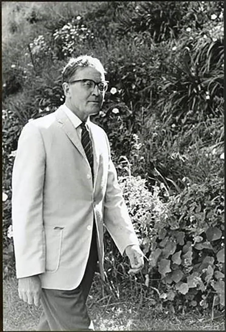 Black and white three-quarter portrait of a man in a suit and wearing glassed in a garden setting.