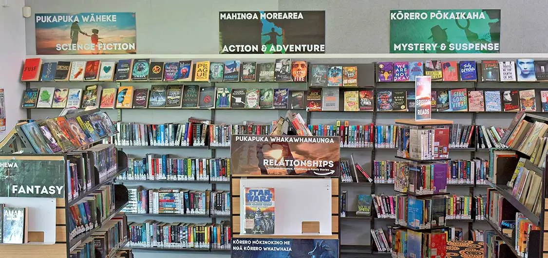 Books arranged by genre inside a school library with signage in te reo Māori and English.