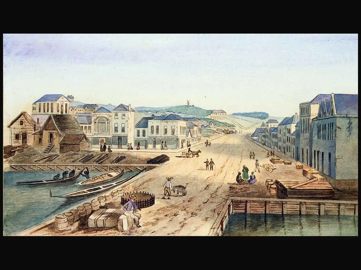 A painting of Queen Street wharf in 1852. Waka (canoes) are drawn up on the shore. On the wharf are barrels, packages, a person sitting on some luggage and another pushing a wheelbarrow. Buildings are along the waterfront and on Queen Street, and a horse is pulling a cart.