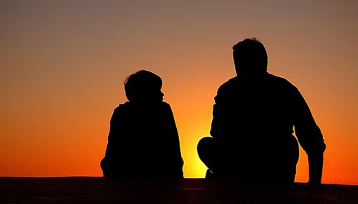 Silhouettes of a father and son sitting and chatting with the sun setting in the background.