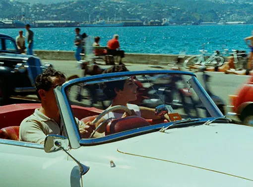 A man and woman driving in an open-top car past the ocean. 