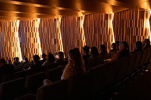 People sitting in a dimly lit auditorium with beautiful decorative wood panels highlighted.