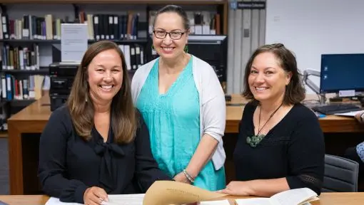 Three women, smiling, while consulting documents in a library reading room setting.