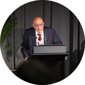 An older Māori man talks at a lectern. He is wearing a suit and has a magnificent bone carving around his neck.  