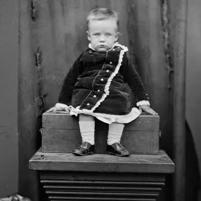 Black and white photo of a small child wearing a formal velvet-looking coat sitting on a plinth. The child looks unimpressed. 