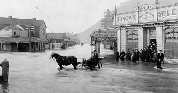 Flooded town street, a horse and cart stands in the floodwaters, and people are grouped outside a building. 