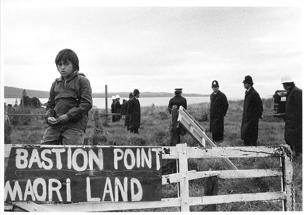 Black and white photograph of the Bastion Point protest, showing a Māori boy beside a sign 'Bastion Point Maori land', with police standing behind him.