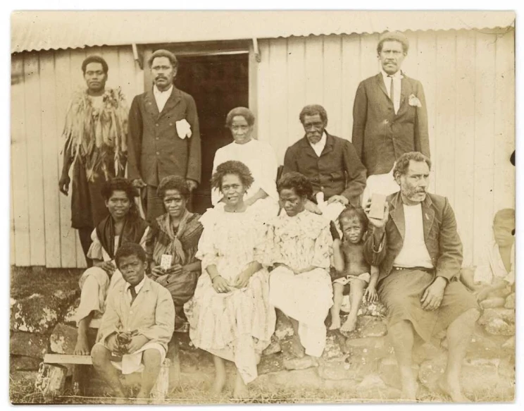 A faded, sepia-toned family portrait of a group out in front of a hut, with the men standing and women and children seated or sitting on the ground.