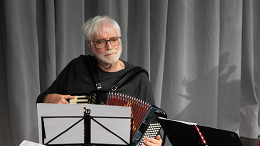 A man plays the accordion on stage while reading from a sheet of music. 