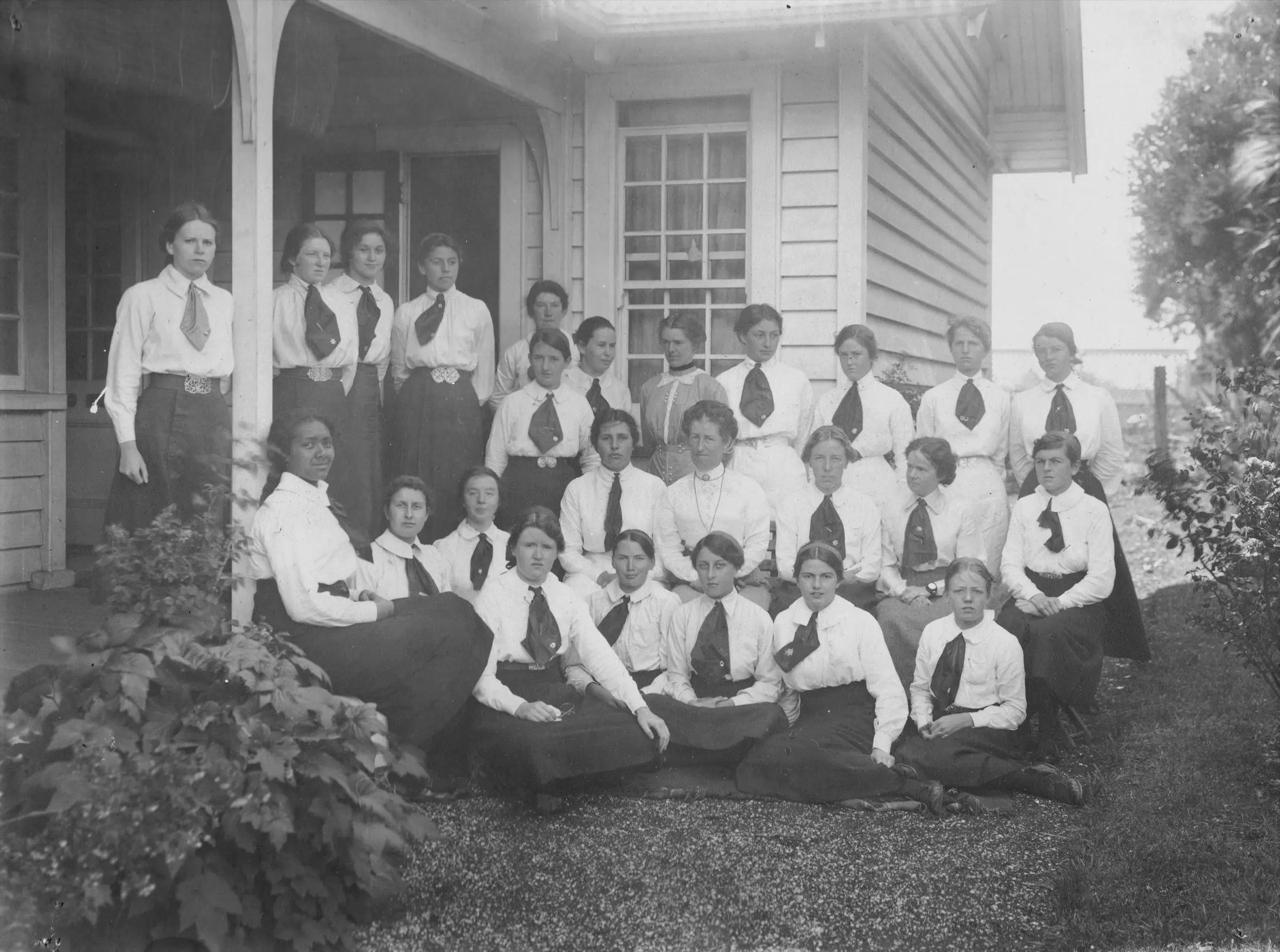 A black and white school portrait showing approximately 20 young women wearing school uniforms and standing or seated out front of a wooden house. 