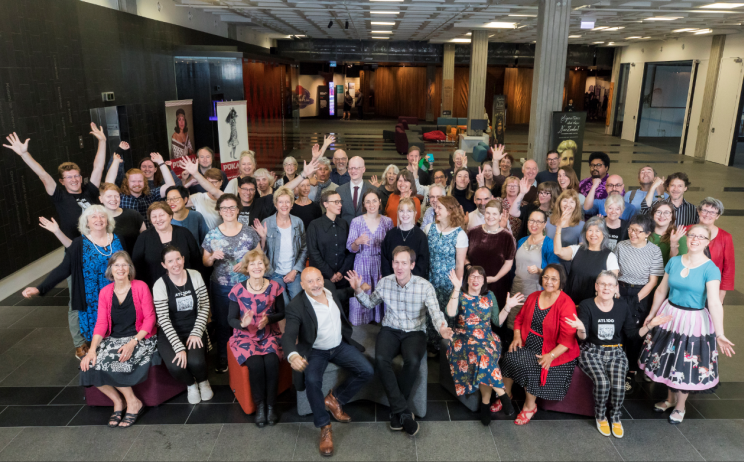 Group portrait of Alexander Turnbull Library staff many holding their hands up and smiling for the camera.