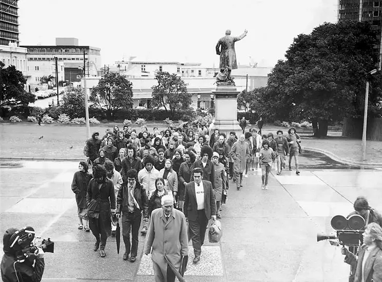 Black and white photo of a large group of Māori people walking towards Parliament led by a kaumatua. They are being filmed. 
