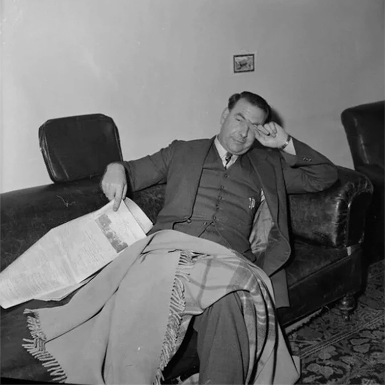 A tired-looking man wearing a three-piece suit reclines on a leather sofa with a rug and newspaper.